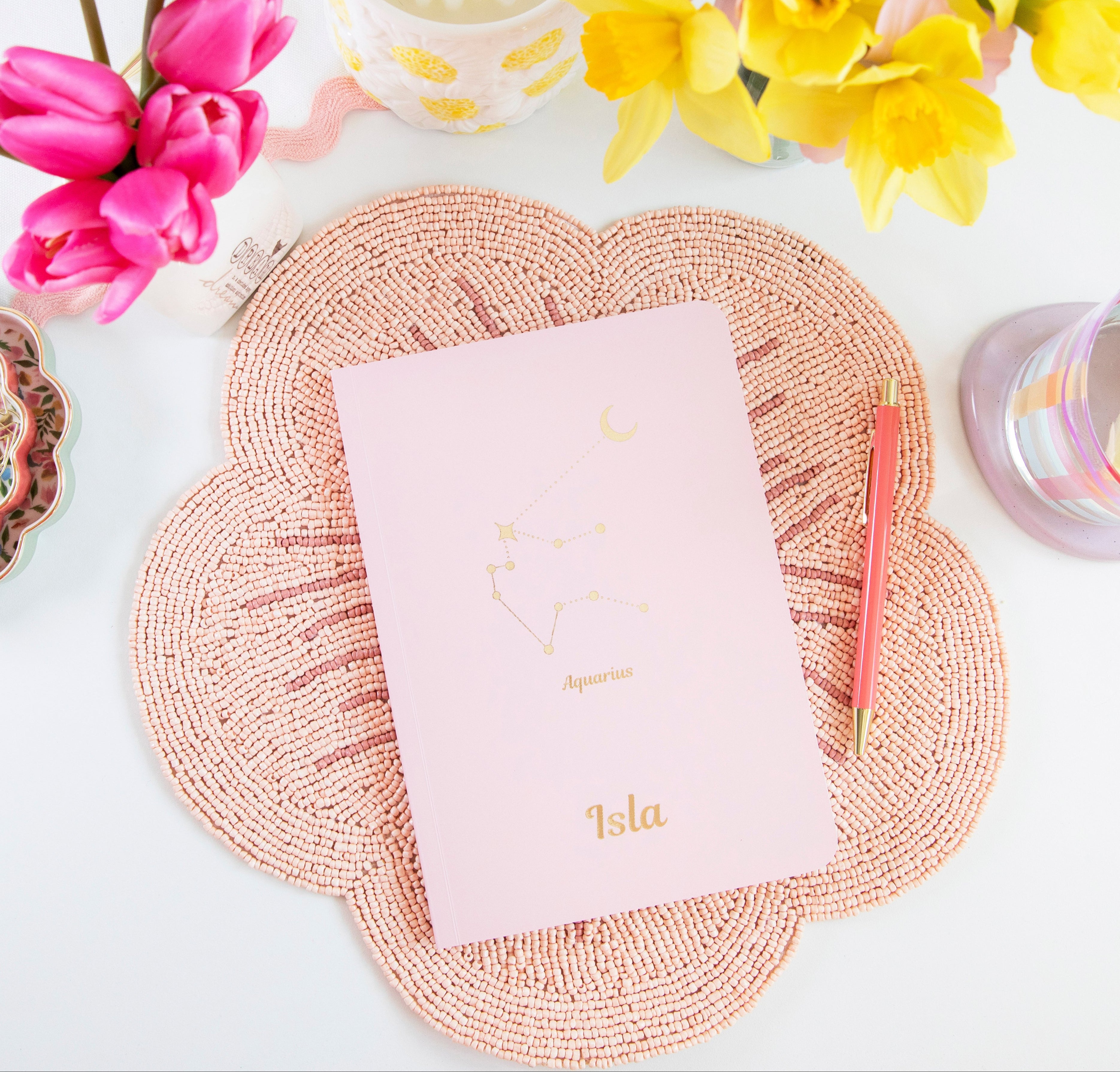 Light pink notebook laid out on flower shapes plate mat, notebook is personalised in gold foil. It says Aquarius star sign and the name Isla underneath